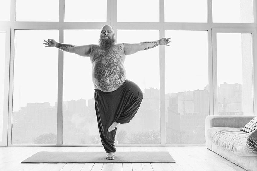 Dickbäuchiger Mann mit Vollbart und tätowiertem Bauch macht Yoga